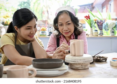 Cheerful Aged Asian Female Potter Teaching A Young Woman To Drawing And Colouring Beautiful Art On The Ceramic Cup In The Workshop.