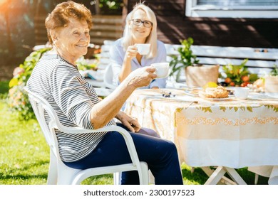 Cheerful aged senior woman drinking tea, sitting outdoor, talking to her adult daughter in rural country house garden on sunny autumn day. Spending quality time with loved ones. - Powered by Shutterstock