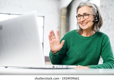 Cheerful aged woman with headset, has wrinkles on the face, dressed in green warm sweater, smiling, at the computer, working online, on the isolation, greeting by waving the hand, happy to see