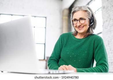 Cheerful aged woman with headset, wrinkles on the face, smiling, listening at the computer, consulting the client online , gives the advice, home working, positive person, takes every chance to work