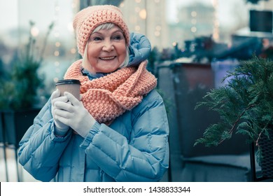 Cheerful aged woman spending time outdoors in a cold winter day and smiling while holding a cup of coffee