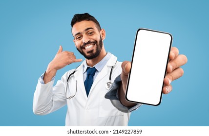 Cheerful Arab Doctor Male Gesturing Call Me, Holding Cell Phone With White Empty Screen, Posing On Blue Studio Background, Smiling To Camera. Medical Consultation By Phone Concept, Mockup