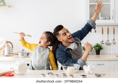 Cheerful arab father and little daughter having fun while baking together in kitchen, happy middle eastern dad and child singing and fooling, using spatula and whisk as microphones, copy space