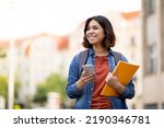 Cheerful Arab Female Student With Smartphone And Workbooks Standing Outdoors, Happy Young Middle Eastern Woman Walking In City After College Classes, Looking Away And Smiling, Copy Space