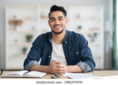 Cheerful arab guy in casual sitting at workdesk and smiling at camera, office interior, copy space. Happy young middle-eastern man sitting at table with notebook and papers, home interior