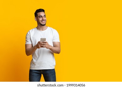 Cheerful arab guy with smartphone looking at copy space and smiling, yellow studio background, panorama. Positive middle-eastern young man chatting on mobile phone, using new application