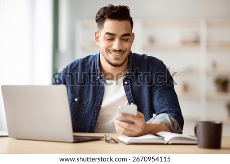 Cheerful arab guy using smartphone while sitting at workdesk