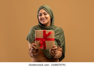 Cheerful Arab Lady In Hijab Holding Gift Box And Smiling To Camera, Positive Muslim Woman With Woman's Day Or Birthday Present Posing Over Beige Studio Background