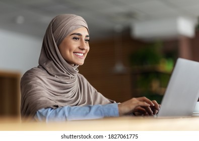Cheerful arab lady in hijab working online at cafe, using laptop, copy space. Side view of smiling muslim woman freelancer typing on laptop, enjoying her new distant job, chatting with clients