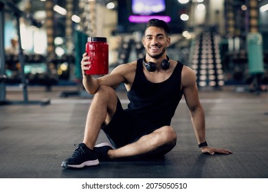 Cheerful Arab Male Athlete Posing At Gym With Container Of Protein Whey In Hand, Smiling Muscular Middle Eastern Man Sitting On Floor In Sport Club And Holding Pack With Fitness Supplements - Powered by Shutterstock