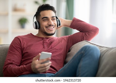 Cheerful arab man listening to music on mobile phone while resting on couch at home, using wireless stereo headset, looking at copy space. Joyful middle-eastern guy enjoying music on smartphone