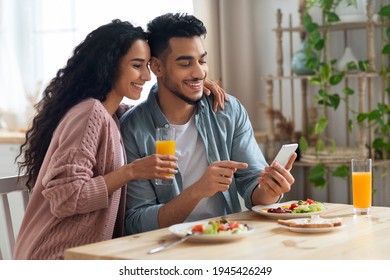 Cheerful Arab Spouses Using Smartphone While Eating Breakfast In Kitchen, Happy Middle Eastern Couple Enjoying Tasty Morning Meal, Shopping Online Or Checking Social Networks On Cellphone, Free Space