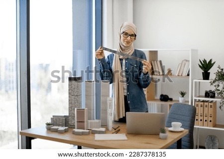 Cheerful arabian female in beige hijab posing with metal ruler behind models of building complex placed near laptop on office desk. Skillful architect using drawing tools for accurate measurements.