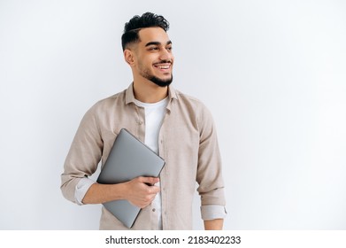 Cheerful arabian or indian young man with beard, wearing casual shirt, standing over isolated white background, holding laptop, looking to the side, smiling joyfully, dreaming, thinking