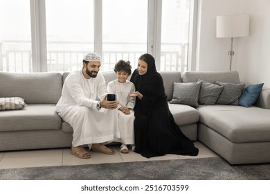 Cheerful Arabian mom, dad and son boy using mobile phone for online communication together, holding cellphone, sitting with gadget on home sofa in comfortable apartment, talking on video call