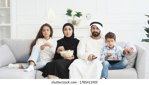 Cheerful Arabian Parents With Little Children Sitting On Couch, Smiling, Watching Movie On TV And Eating Popcorn. Cute Arab Small Kids With Mother And Father Spending Time Together At Home.