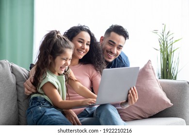 Cheerful Arabic Family Having Fun With Laptop At Home, Watching Photos Together. Middle Eastern Parents And Their Little Daughter Using Computer While Sitting On Couch In Living Room, Copy Space