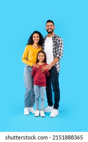 Cheerful Arabic Family Of Three Posing Together, Father And Mother Embracing Their Kid Daughter Standing On Blue Studio Background. Full Length, Vertical Shot