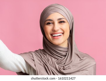 Cheerful arabic girl in hijab taking selfie and sincerely laughing, pink background with free space