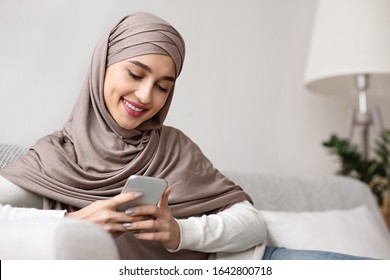 Cheerful arabic girl in hijab using smartphone at home, touching screen, texting while sitting on couch in living room, closeup
