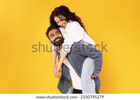 Cheerful Arabic Guy Carrying His Girlfriend On Back, Piggybacking Loving Woman For Fun, Posing And Fooling Together Over Yellow Background. Couple Having Fun, Studio Shot. Love And Happiness