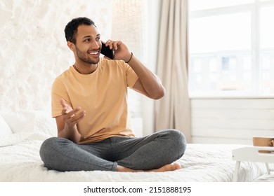 Cheerful Arabic Man Talking On Mobile Phone Communicating Looking Aside Sitting On Bed In Modern Bedroom At Home. Guy Calling Speaking By Cellphone Indoors. Communication Concept