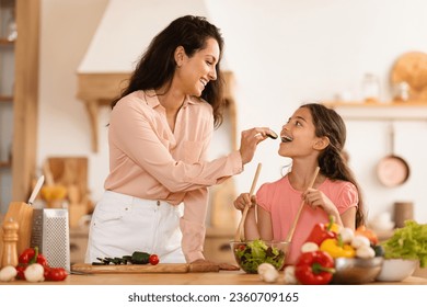 Cheerful Arabic Mommy And Her Daughter Kid Girl Cooking Together, Mom Feeding Preteen Child Giving To Taste Cucumber Slice, Having Fun While Preparing Family Dinner In Kitchen At Home Interior - Powered by Shutterstock