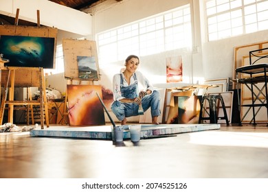 Cheerful artist squatting close to her painting on the floor. Happy female painter smiling at the camera while holding a paintbrush. Creative young woman working on a new painting in her art studio.
