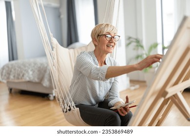 Cheerful artistic senior blonde woman painting and brushes canvas while sitting rope swing in her living room 
