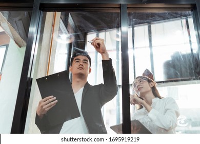 Cheerful Asian Boss And Secretary Holding Clipboard, Writing On Glass Wall With Pen In Modern Office.