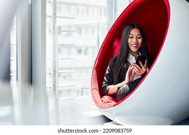 Cheerful Asian Business Woman Using Mobile Phone Sitting Inside Ball Chair At Office 