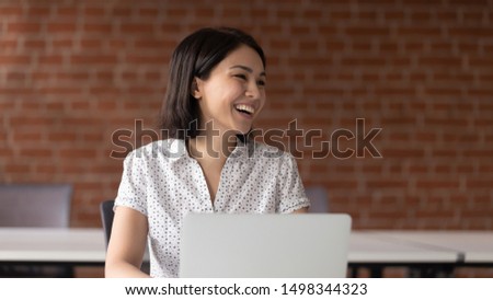 Cheerful asian business woman working with laptop in loft office, having break, talking with somebody aside, laughing, having fun. Happy female employee enjoying pause time, joking with colleagues.