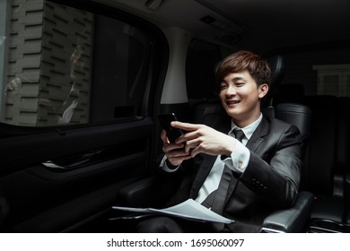 Cheerful Asian Businessman On Black Suit Using Smartphone And Laughing In Luxury Car.