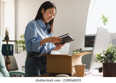 Cheerful Asian Businesswoman Unpacking Box Holding Book Standing In Modern Office Indoors. Business Literature, Shopping And Delivery Service Concept. Side View - Powered by Shutterstock