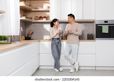 Cheerful asian couple enjoying morning coffee at home, happy pretty young lady having conversation with her husband, standing by kitchen table, holding mugs and chatting, full length, copy space - Powered by Shutterstock