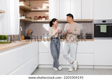 Cheerful asian couple enjoying morning coffee at home, happy pretty young lady having conversation with her husband, standing by kitchen table, holding mugs and chatting, full length, copy space