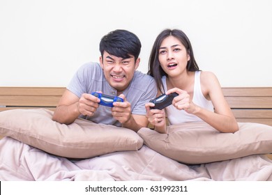 Cheerful Asian Couple In Love Playing Video Games With Joysticks On The Bed