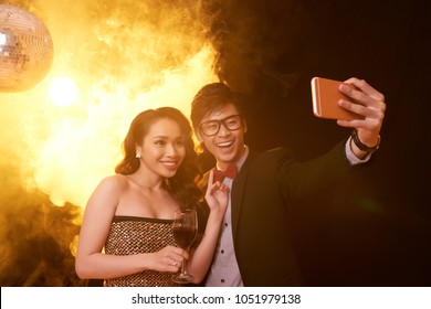 Cheerful Asian Couple Taking Selfie On Smartphone While Enjoying Party At Night Club, Pretty Woman Holding Glass Of Red Wine In Hand