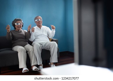 Cheerful Asian Elderly Couple in 3D glasses while sitting on couch watching virtual cinemas or movies over blue wall background at their house. Having Fun, Enjoy in Retirement life. - Powered by Shutterstock