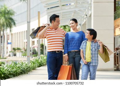 Cheerful Asian Family Of Three Holding Paper Bags In Hands And Sharing Emotions With Each Other After Completion Of Productive Shopping, Facade Of Modern Shopping Mall On Background