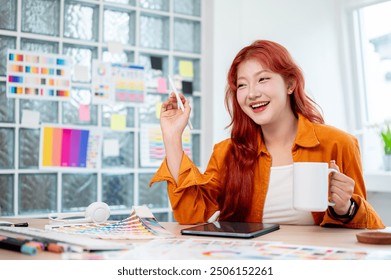 A cheerful Asian female graphic designer with red curly hair is smiling at the camera, holding a coffee mug and sitting at her desk in the office, surrounded by color swatch palettes. - Powered by Shutterstock