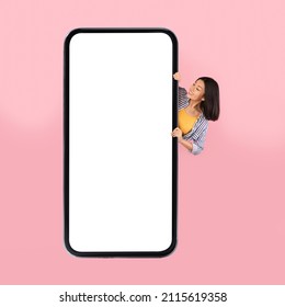 Cheerful Asian Female Hiding Behind Big Cellphone And Looking At Huge Blank Phone Screen Over Pink Studio Background. Great Mobile Offer Concept. Mockup, Square Shot
