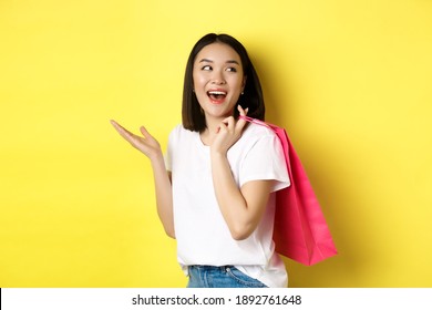 Cheerful asian female shopper looking amused, holding shopping bag and looking right, standing over yellow background