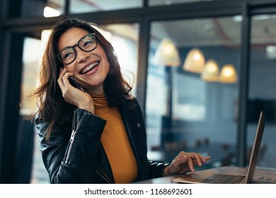 Cheerful asian female talking on mobile phone while sitting on desk with laptop. Business woman in casuals making a phone call and laughing. - Powered by Shutterstock