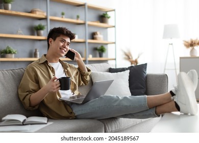 Cheerful Asian Freelancer Guy Drinking Coffee And Talking On Cellphone While Having Break During Work With Laptop Computer At Home Office, Speaking With Friends, Resting Sitting On Sofa, Free Space