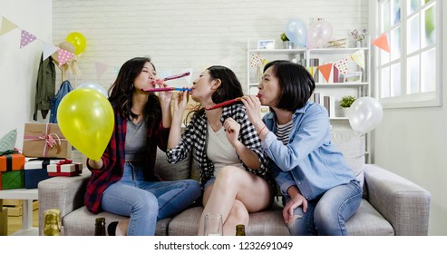 Cheerful Asian Friends Looking Blowing Blowers Having Fun. Young Ladies Holding Whistles Playing At House Birthday Party In The Daytime In Living Room. Decorated Home Full Of Gift Box Balloons Flags.