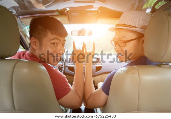 Cheerful Asian friends posing for photography and\
showing sign of horns while sitting in car ready for unforgettable\
road trip, lens flare