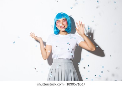 Cheerful Asian Girl In Blue Wig Throwing Confetti Over White Background, Celebrating Halloween, Waving Hand To Say Hello, Greeting Someone