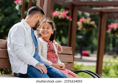 Cheerful Asian Girl Looking At Tattooed Dad On Bench In Park