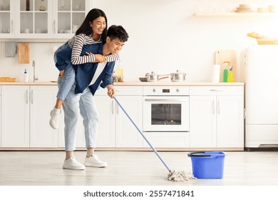 Cheerful asian guy piggyback his pretty girlfriend while mopping floor at cozy white kitchen, panorama with copy space. Loving korean couple having fun while house-keeping their new apartment - Powered by Shutterstock